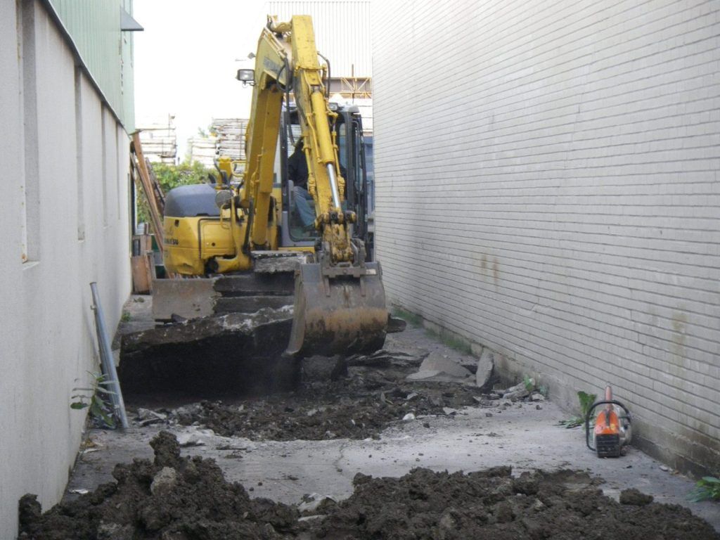 Pelle hydraulique cassant une dalle de béton dans une allée étroite entre deux bâtiments